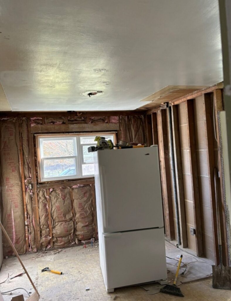 A kitchen under renovation with exposed wall insulation, an unfinished ceiling, and a refrigerator in the middle of the room. Tools are scattered on the floor.