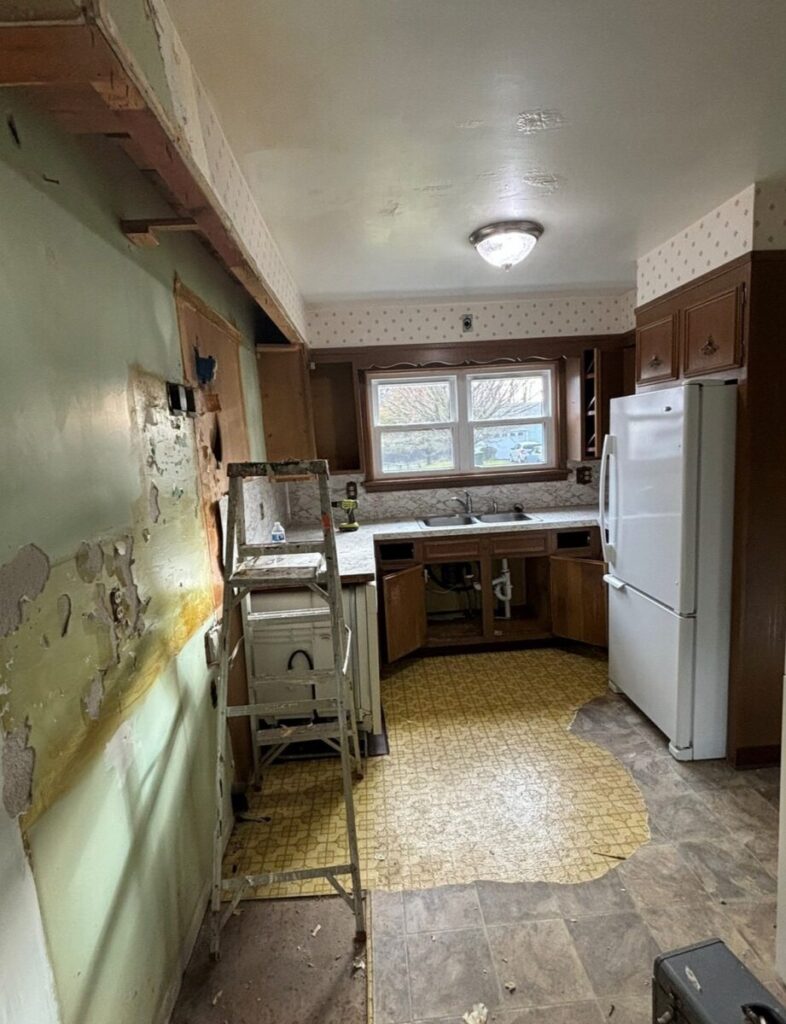 A kitchen under renovation with partially stripped walls, exposed drywall, and missing cabinets. A ladder and various tools are scattered around. The floor has sections of old and new tiles.