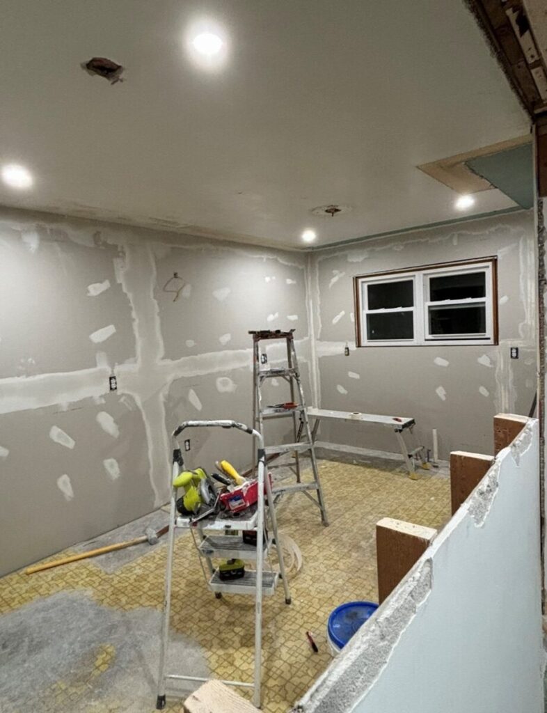 A partially renovated room with ceiling lights, patched walls, and construction tools. A step ladder is in the center, and various tools are scattered on the floor. A window is visible on the far wall.