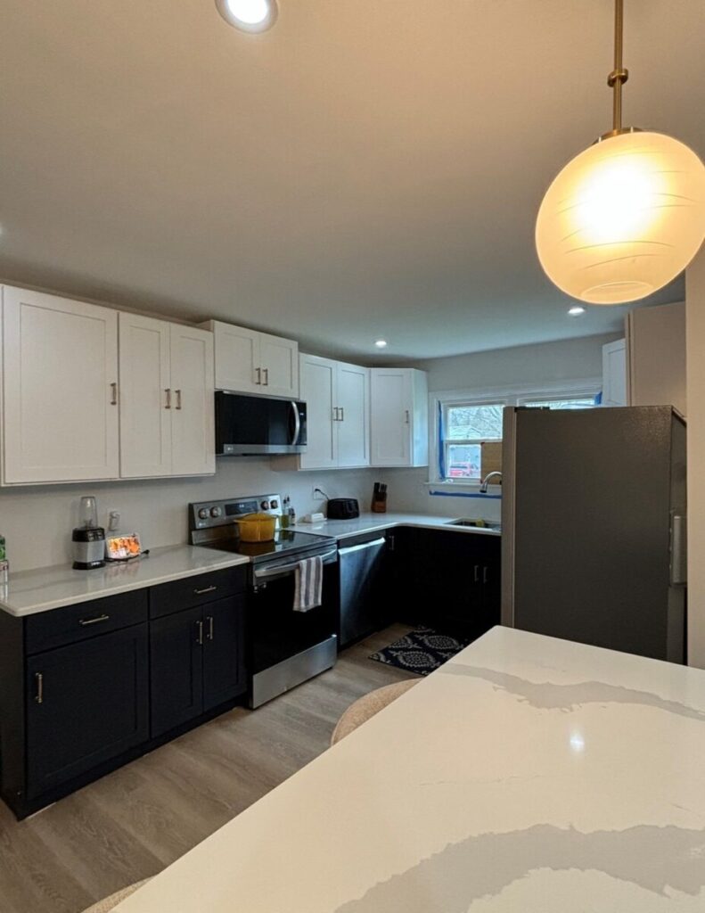 Modern kitchen with black lower cabinets and white upper cabinets. Stainless steel appliances, a large refrigerator, and a marble countertop island are present under soft overhead lighting.