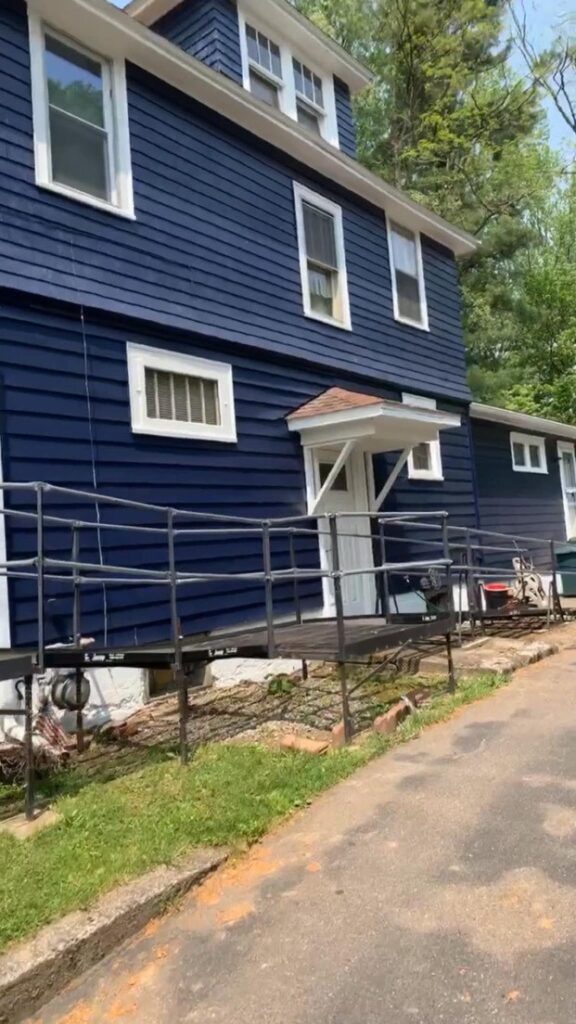 A blue two-story house with white trim and a small porch over the front door. A ramp with a metal railing leads to the entrance. Trees and a driveway are in the background.