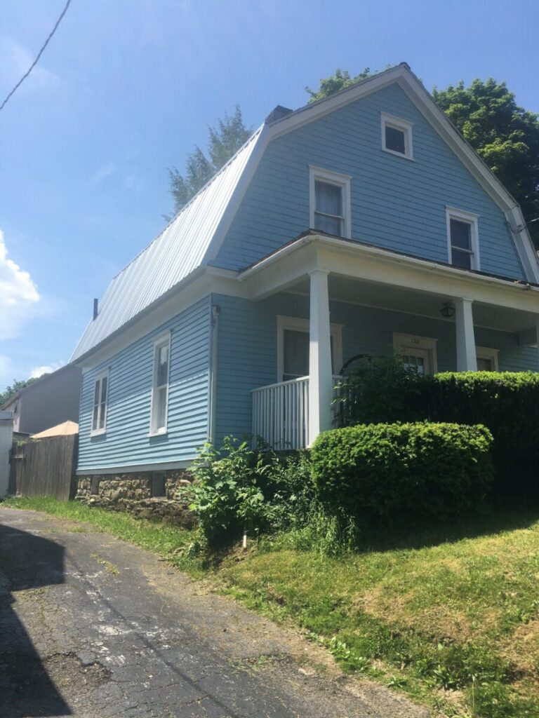 A two-story blue house with a white porch, surrounded by green bushes and trees, and a paved driveway on a sunny day.