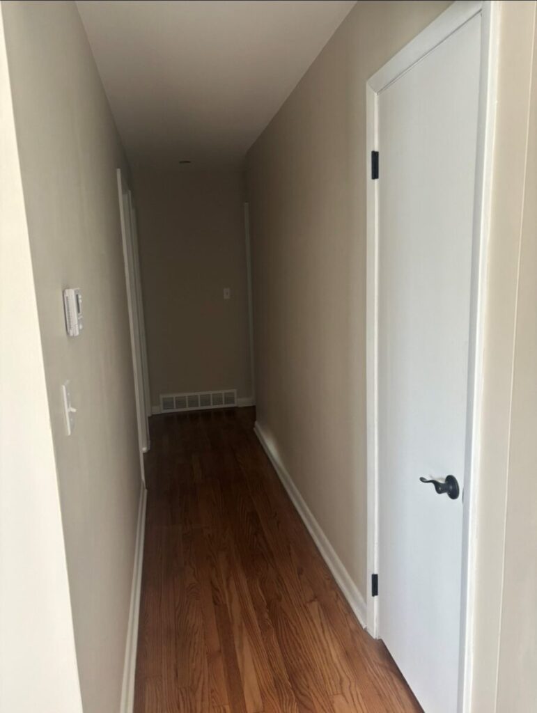 A narrow hallway with wooden flooring, beige walls, and white doors is shown. A thermostat is mounted on the left wall. The hallway appears clean and well-lit.